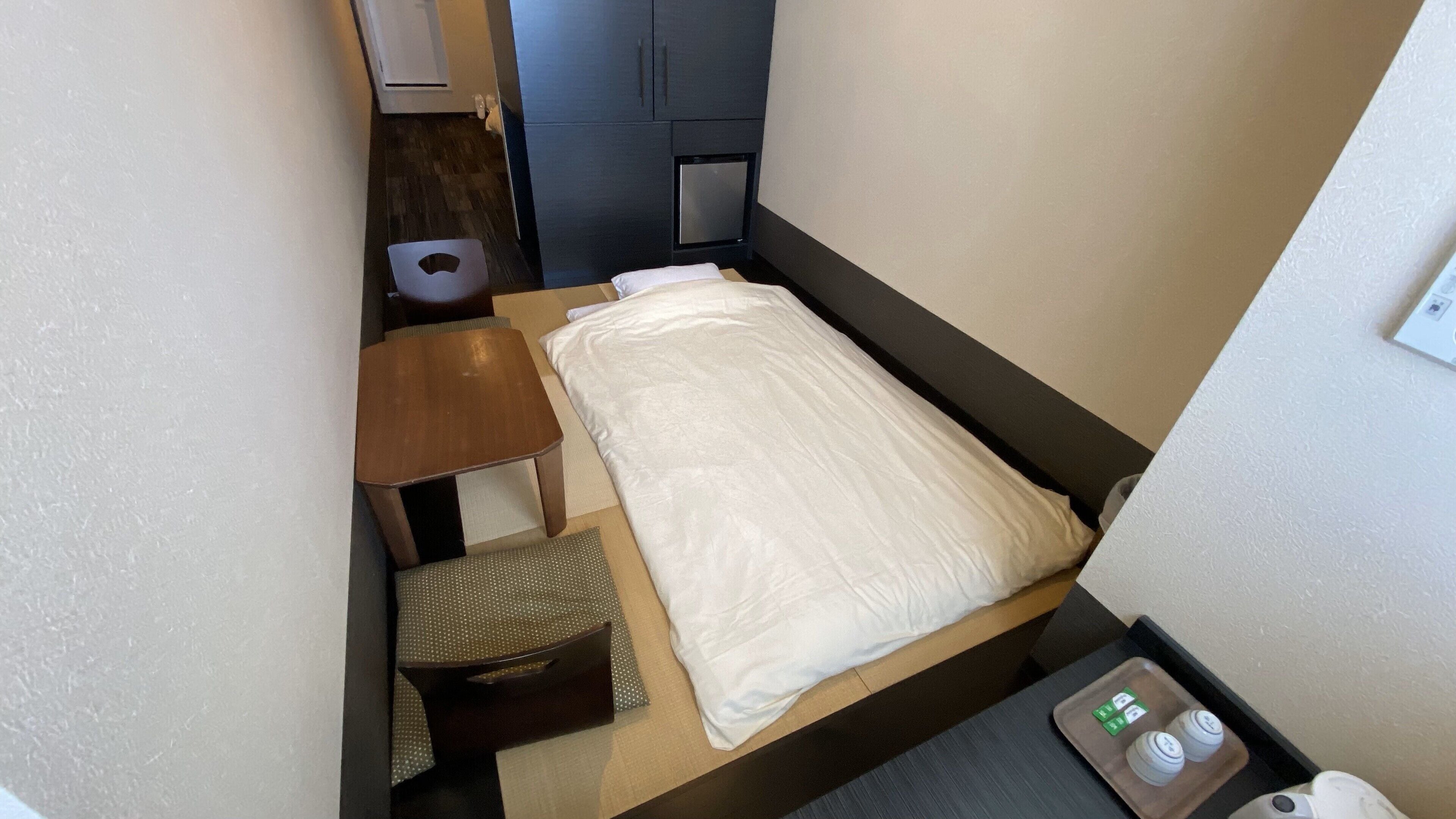 Habitación de hotel pequeña y funcional con una cama individual, una mesita de noche de madera, una pequeña mesa y una silla. Hay un mueble bajo y un calentador en el suelo.