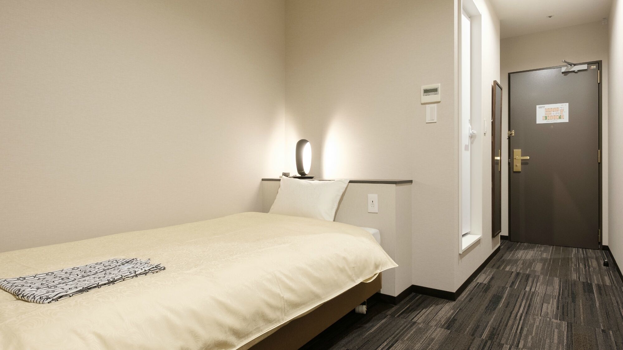 A hotel room with a bed, a nightstand, a lamp, a door, and a carpeted floor.