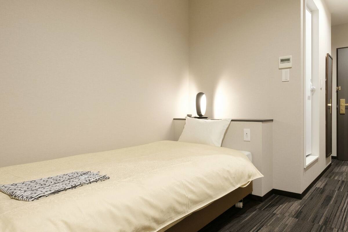 A hotel room with a bed, a nightstand, a lamp, a door, and a carpeted floor.