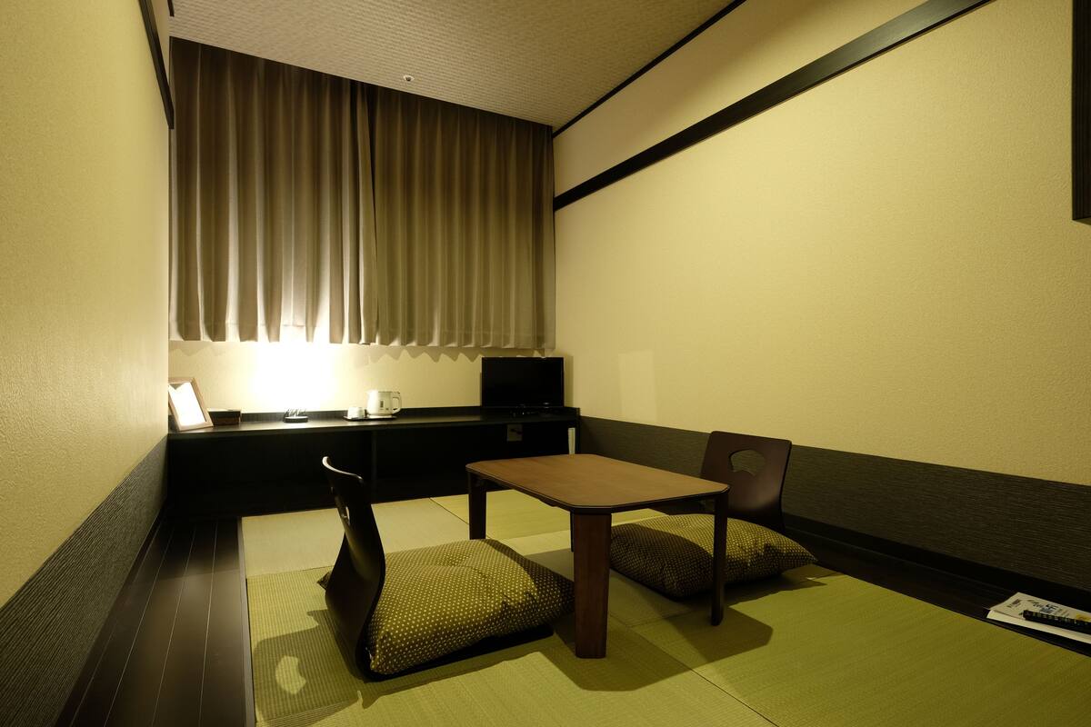 A small, dimly lit room with a single bed, a bedside table, a chair, and a television.