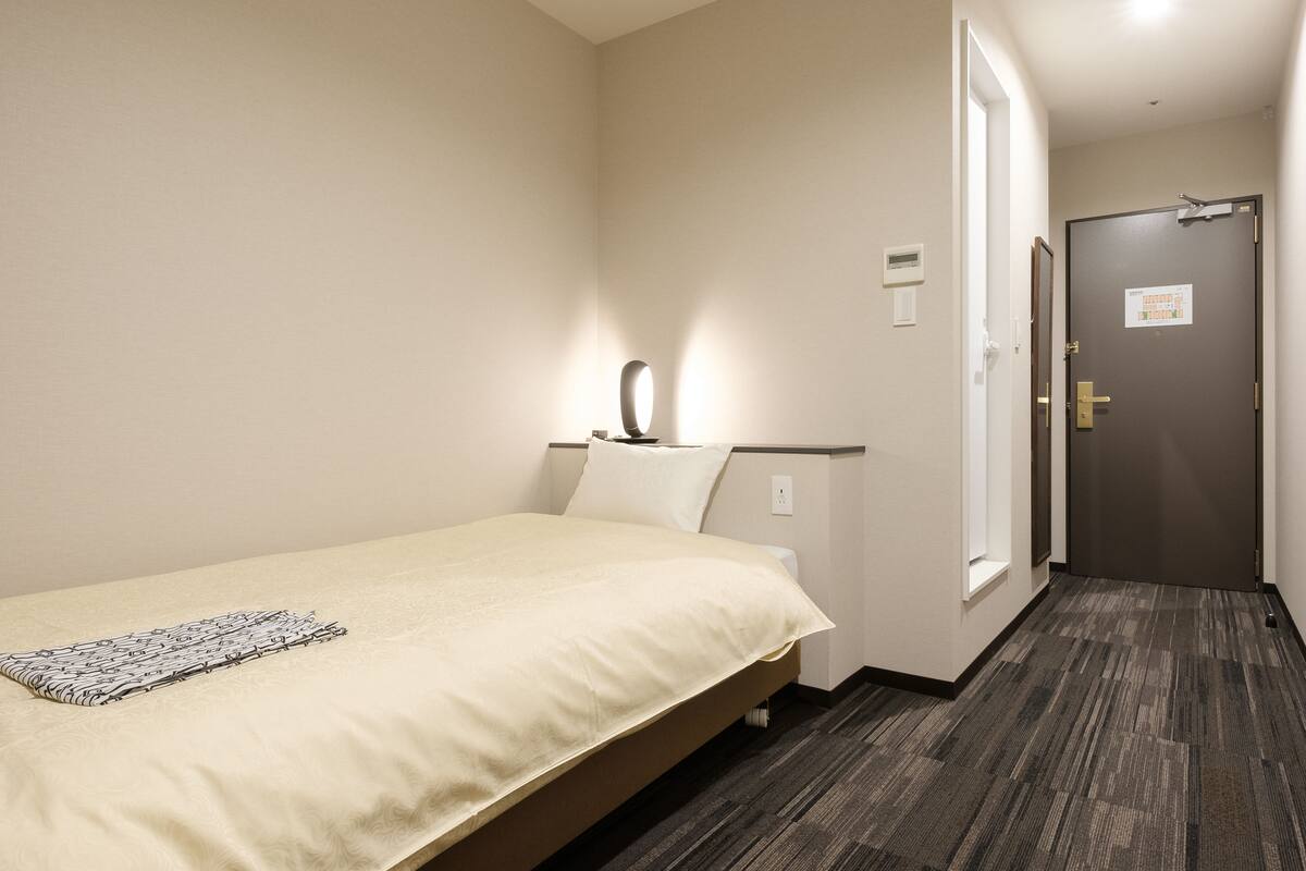 A hotel room with a bed, a bedside table, a lamp, and a door.