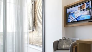 Une chambre d’hôtel moderne avec une grande fenêtre, un téléviseur fixé au mur, un fauteuil gris, une table en bois et un sac à main gris posé sur le fauteuil.