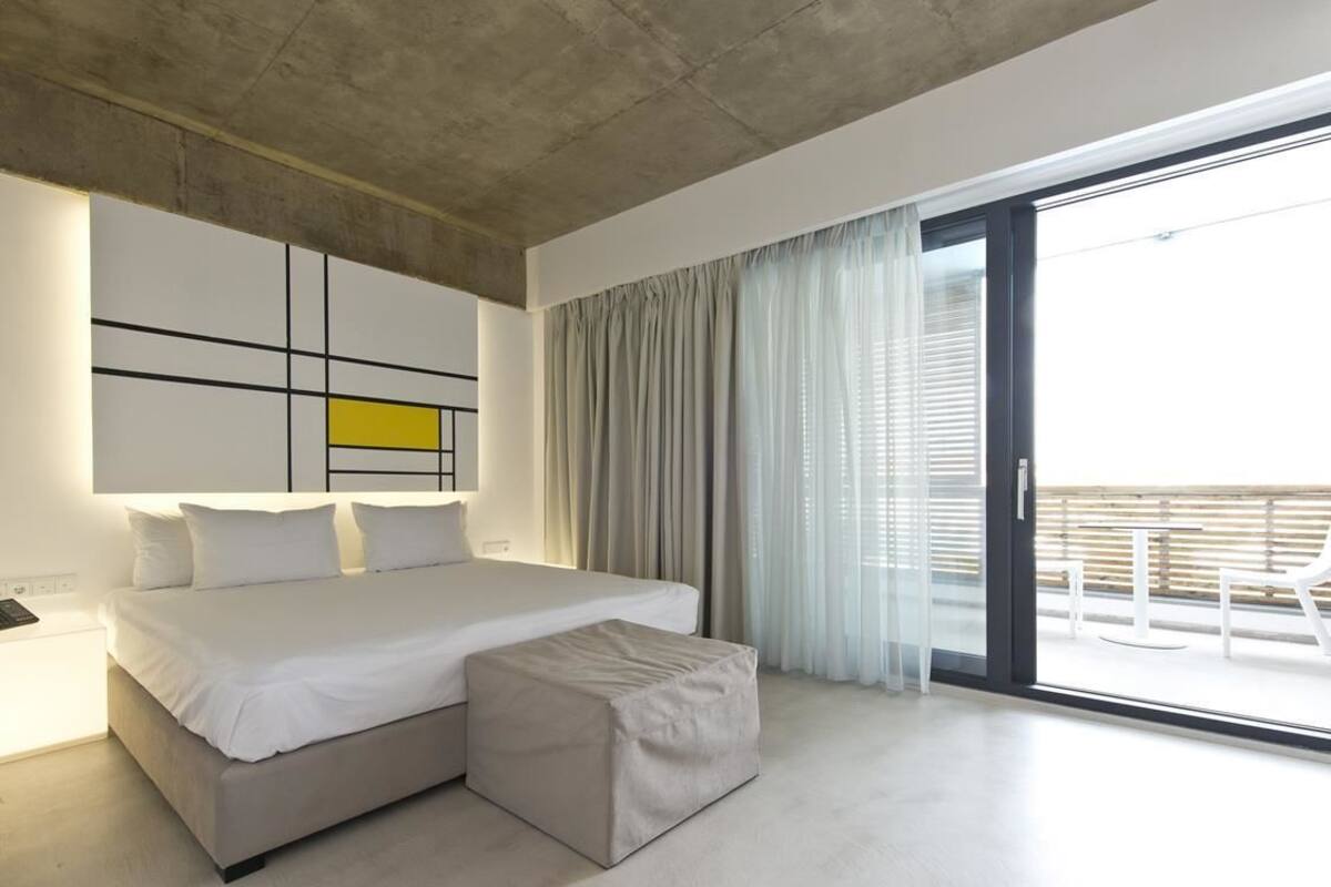 Un dormitorio moderno con una cama grande, un pequeño reposapiés, una obra de arte minimalista en la pared y una puerta corrediza de vidrio que da a un balcón.