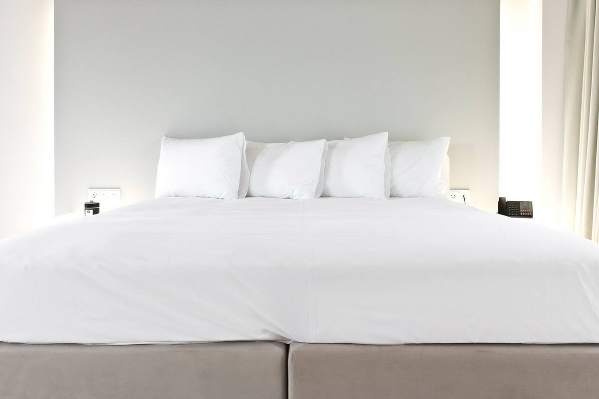 Una cama bien hecha con sábanas blancas, un cabecero, una mesita de noche con una lámpara y un teléfono a la derecha.