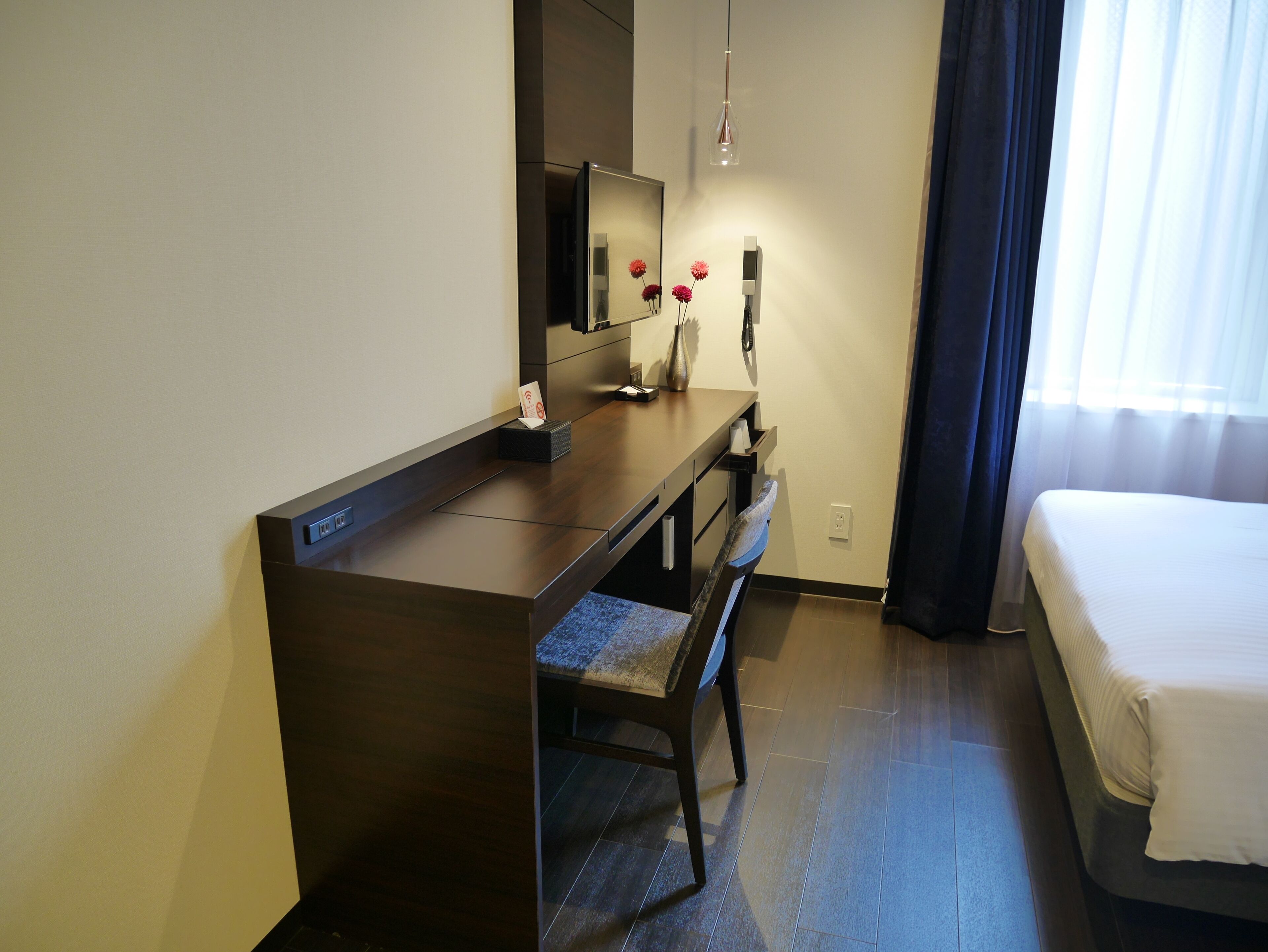 A hotel room with a wooden desk, a chair, a bed, a mirror, and a vase with flowers.