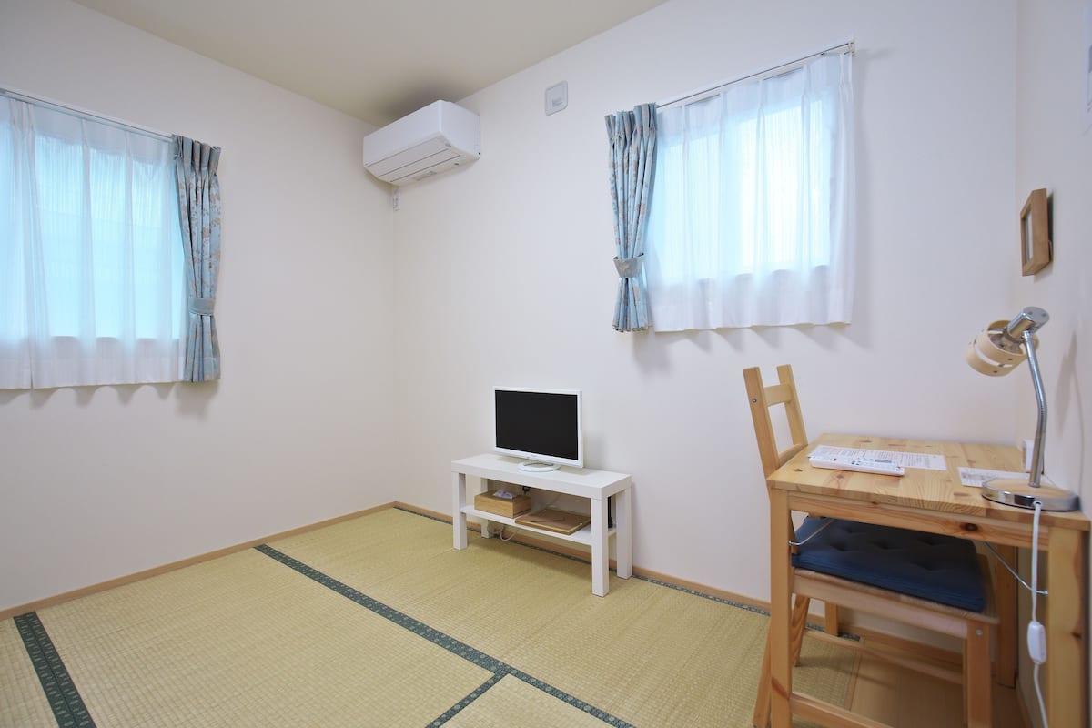 A room with a TV on a white stand, a desk with a lamp, and a chair.