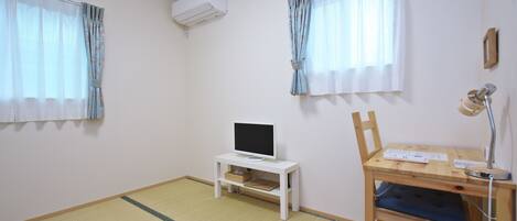 A room with a TV on a white stand, a desk with a lamp, and a chair.