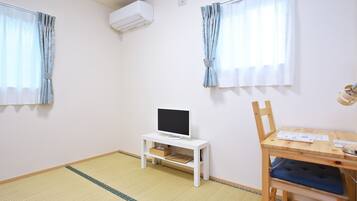 A room with a TV on a white stand, a wooden desk with a chair, and a window with curtains.