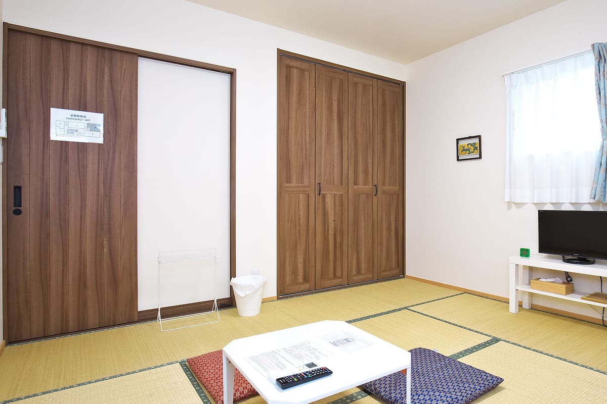 A room with a tatami mat floor, a television on a white stand, a wooden wardrobe, and a small table with papers and a remote control.