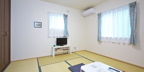 Una habitación con suelo de esteras de tatami, un televisor sobre un pedestal blanco y dos ventanas con cortinas.