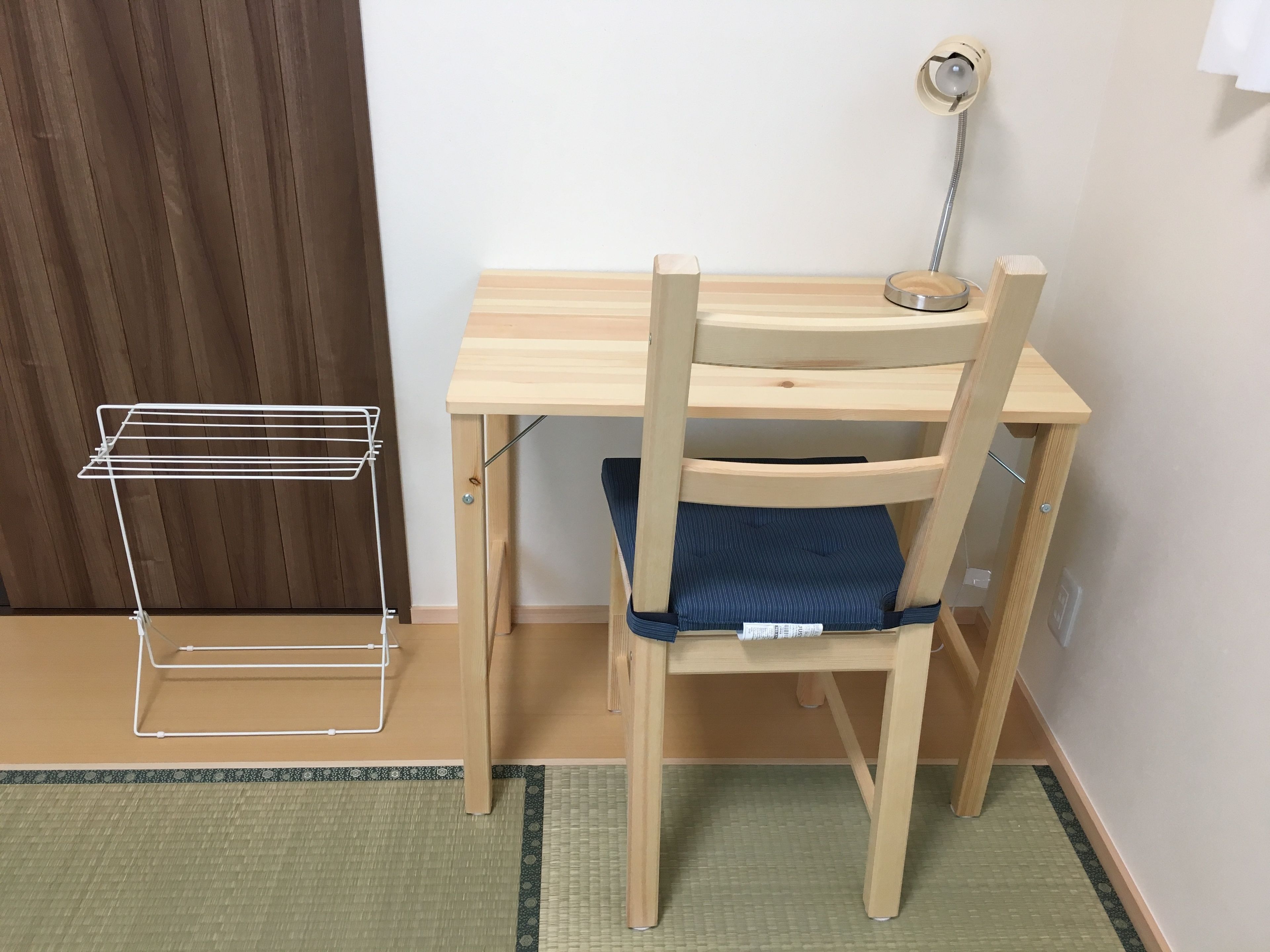 A wooden desk with a chair, a white wire shelf, and a floor lamp.