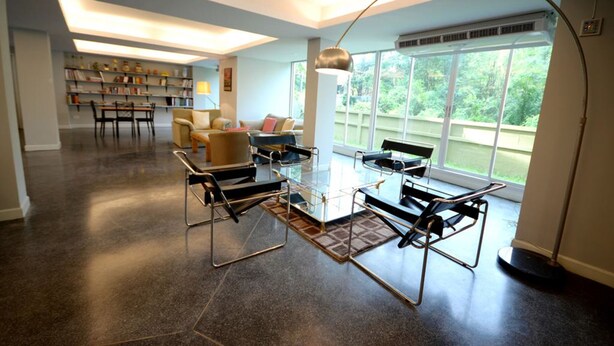 Een ruime woonkamer met een glazen salontafel, moderne fauteuils, een boekenkast en een groot raam met uitzicht op groen.