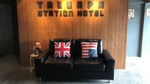 Un área de recepción con una pared de madera que muestra el nombre 'TALUAPA STATION HOTEL'. Hay un sofá de cuero negro con dos almohadones, uno con la bandera del Reino Unido y el otro con la bandera de los Estados Unidos. A la izquierda, una pequeña mesa con un jarrón de flores.