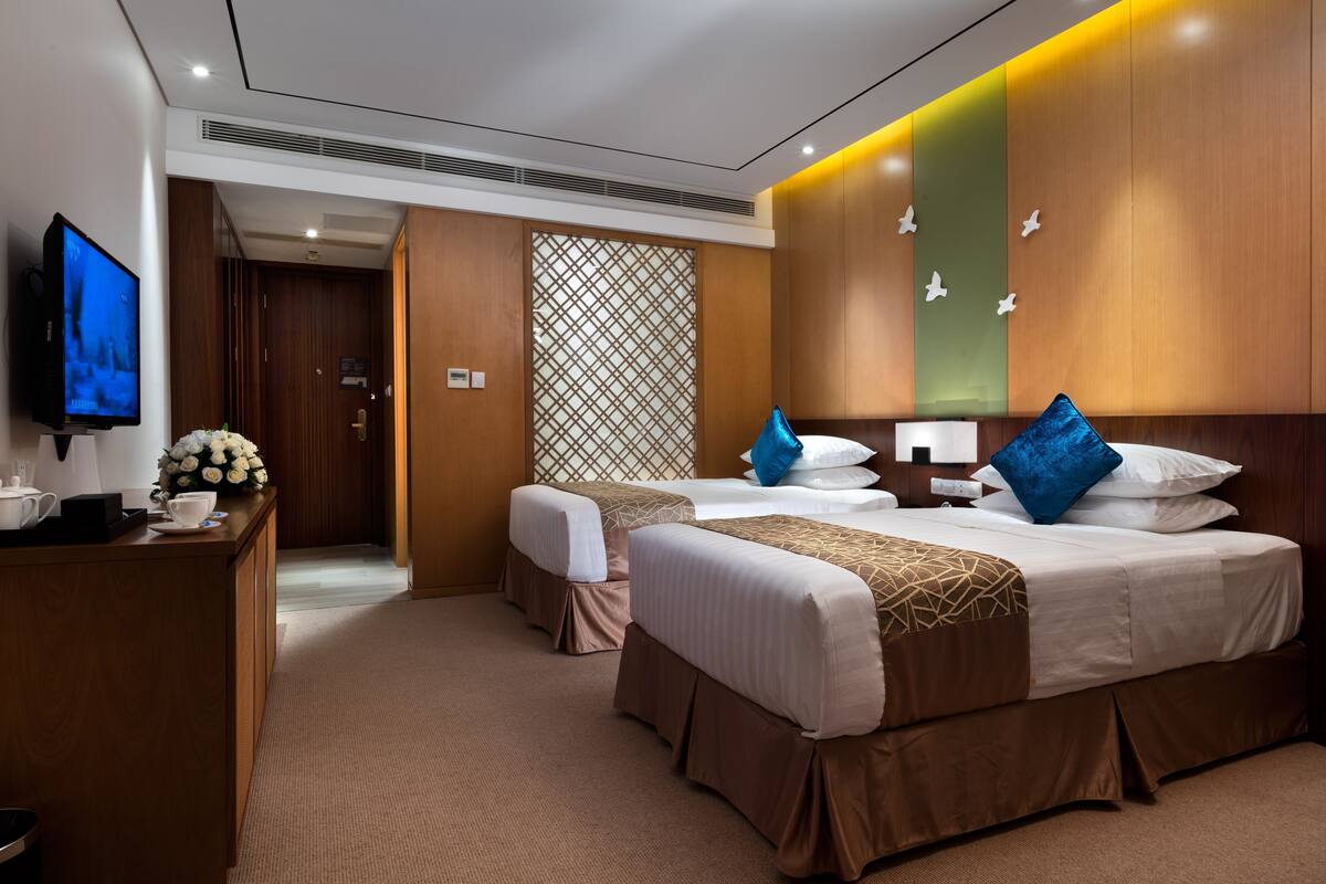 A hotel room with two beds, a TV, a desk with a flower arrangement, and a decorative wall.