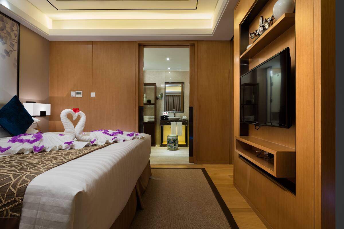 A hotel room with a bed, a television, a bathroom, and a hallway leading to a kitchen.