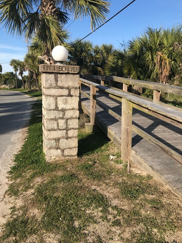 A stone sign with "FORT SCREWELL" engraved, a wooden boardwalk, and palm trees in the background.