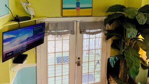 Ein Zimmer mit gelben Wänden, einem Fernseher auf einem Schrank, einer großen Pflanze und einer Tür, die nach draußen führt.