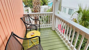 Ein überdachter Balkon mit einem gelben Tisch und zwei Netzstühlen, einem weißen Geländer und Blick auf benachbarte Häuser.