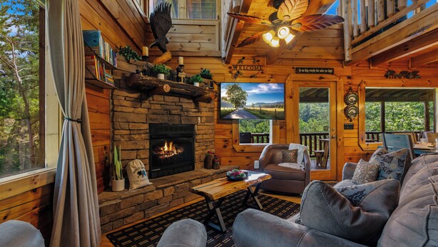 A cozy cabin interior with a fireplace, wooden walls, a sofa, and a view of the outdoors.