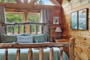 A log cabin interior with a bed, curtains, a lamp, and framed artwork.