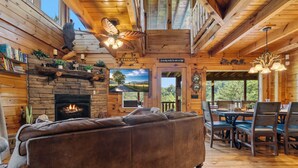 A cozy cabin interior with a fireplace, a large sofa, a dining area, and a view of the outdoors.