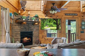 L’intérieur chaleureux d’un chalet avec une cheminée en pierre, des murs en bois, un coin salon confortable et un téléviseur fixé au mur.
