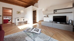 A spacious living room with a wooden ceiling, a large flat-screen TV, a glass coffee table with a book, and a modern kitchen in the background. - EXCLUSIVE AND ROMANTIC CHARME APARTMENT IN THE CORE OF HISTORIC CENTER OF MODICA (Modica)
