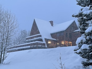 A large, multi-story lodge with a steeply pitched roof covered in snow, surrounded by snow-laden trees.