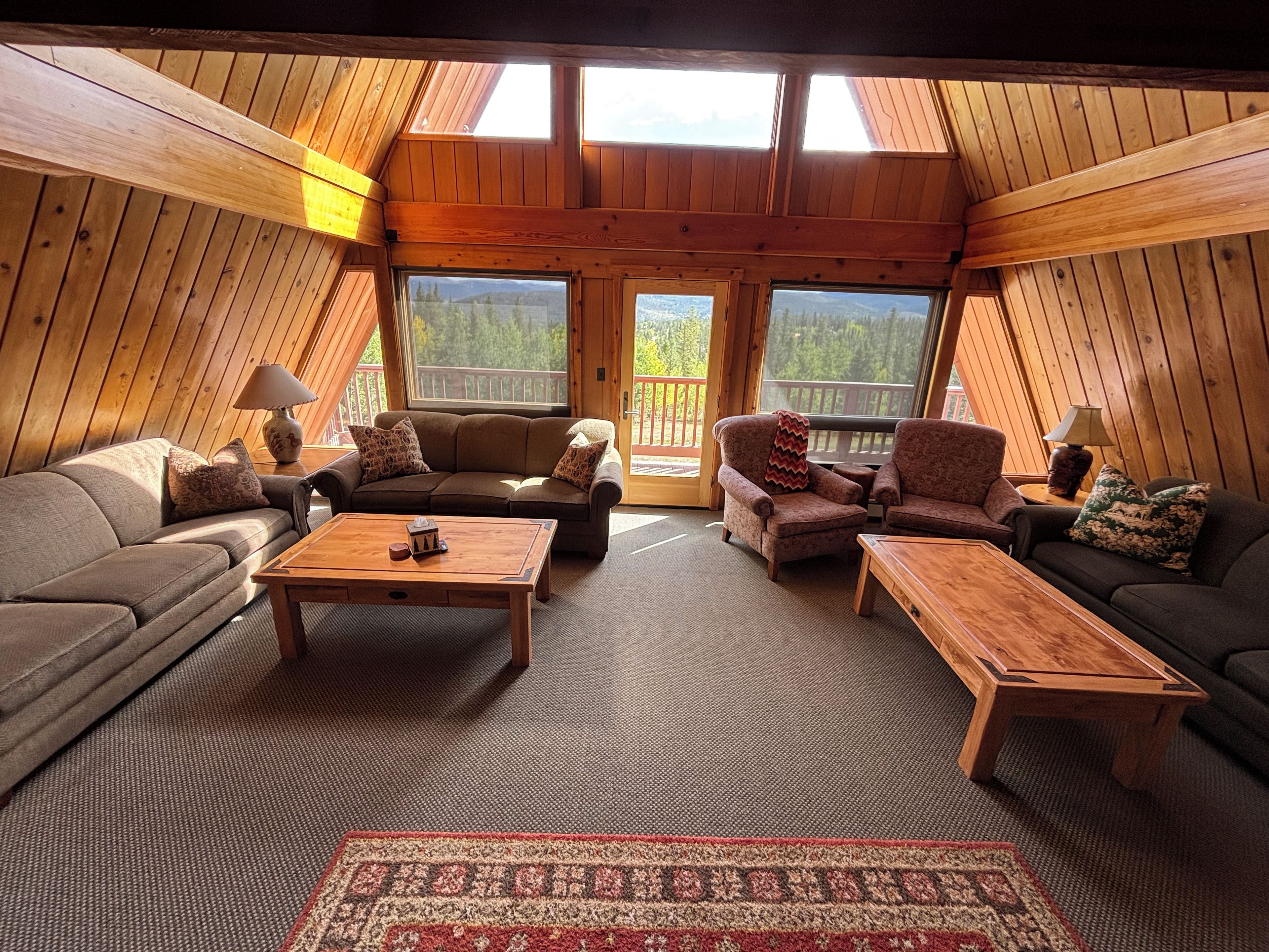 A cozy loft-style room with a high ceiling, wooden walls, and large windows offering a view of the outdoors.