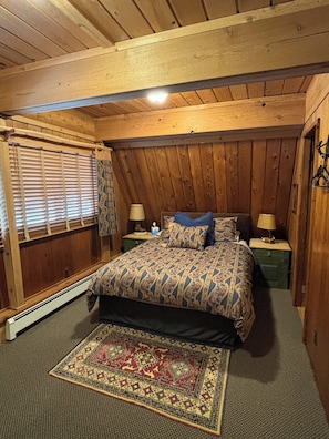 A bedroom with a wooden ceiling, a bed with patterned bedding, two bedside tables with lamps, a window with blinds, and a rug on the floor.