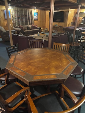 A wooden octagonal table surrounded by chairs in a room with wooden walls and a television mounted on the wall.