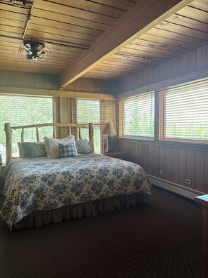 Une chambre avec un plafond en bois, un lit recouvert d’une couvre-lit à motifs floraux et des fenêtres équipées de stores.