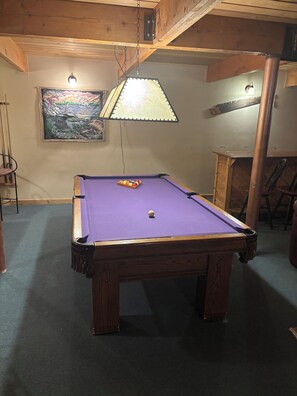 Une table de billard avec une bille au centre, une lampe au-dessus et un tableau accroché au mur.