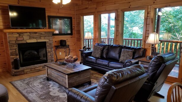 A cozy cabin-style living room with a stone fireplace, leather furniture, and large windows offering a view of the outdoors.