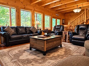 A cozy log cabin interior with a leather sofa, a wooden coffee table, and a large window with a view of trees. - Free Daily Activities- Enjoy family time in our theater game room or hot tub (Sevierville)