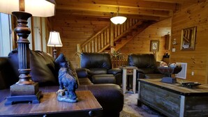 A cozy log cabin interior with leather sofas, a wooden coffee table, and a staircase leading to the upper level.