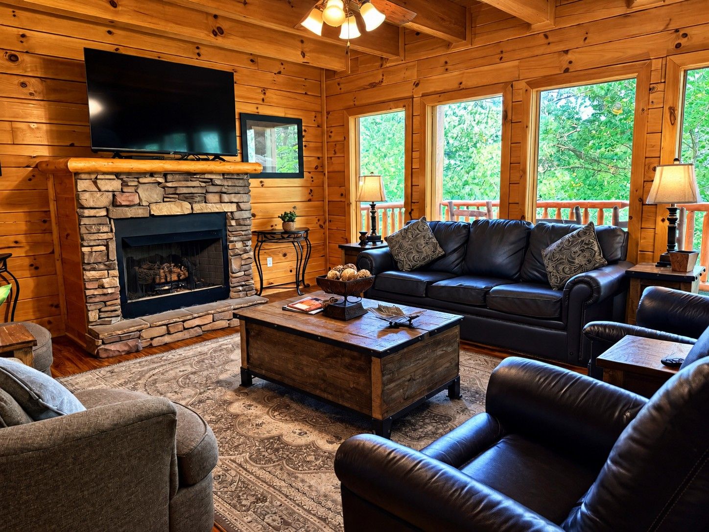 A cozy log cabin interior with a stone fireplace, leather furniture, and large windows.