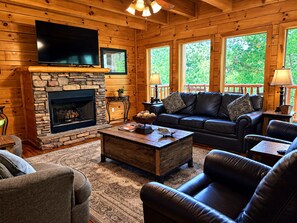 A cozy log cabin interior with a stone fireplace, leather furniture, and large windows.