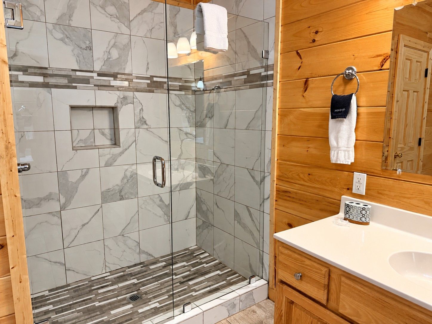 Un baño moderno con ducha envuelta en vidrio, baldosas de mármol, muebles de madera y un lavabo blanco.