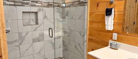 Un baño moderno con ducha envuelta en vidrio, baldosas de mármol, muebles de madera y un lavabo blanco.