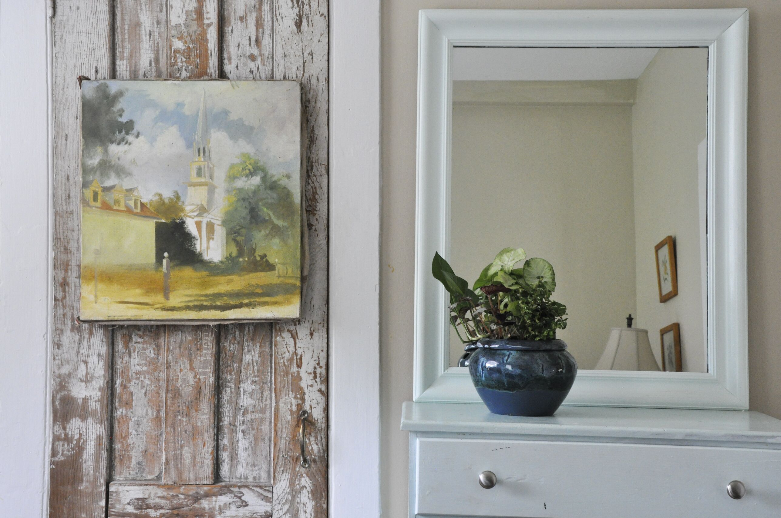 A rustic wall with a weathered door and a painting of a church. A blue vase with a plant is on a white dresser next to a mirror reflecting a lamp and picture frames.