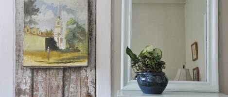 A rustic wall with a weathered door and a painting of a church. A blue vase with a plant is on a white dresser next to a mirror reflecting a lamp and picture frames.