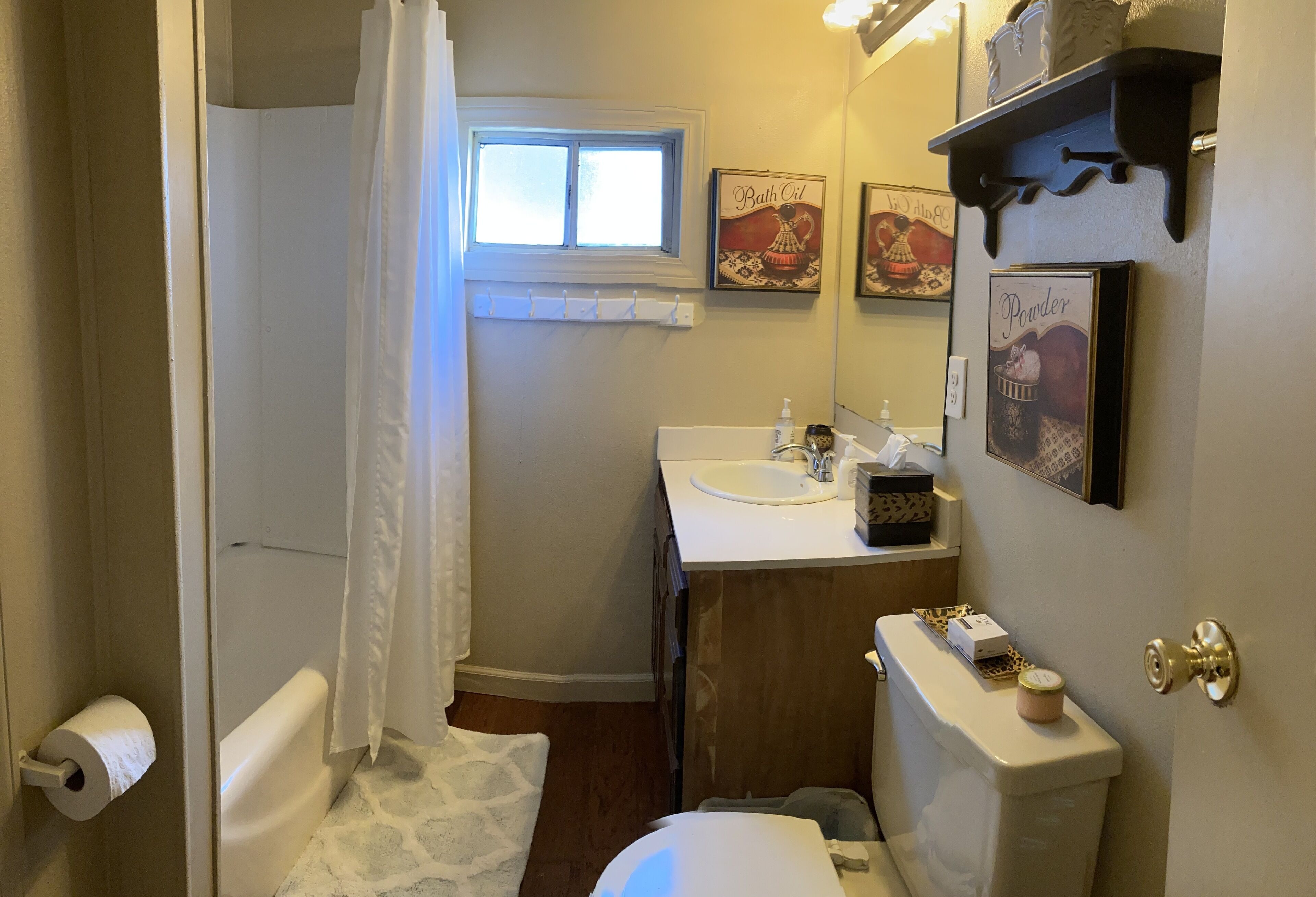 Ein Badezimmer mit Toilette, Waschbecken und Duschvorhang. An der Wand befinden sich Bilder in Rahmen, und ein Ablagefach enthält Pflegeprodukte.