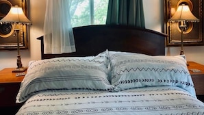 Un dormitorio con una cama de madera oscura, ropa de cama blanca y estampada, dos lámparas de noche y una ventana con cortinas verdes.