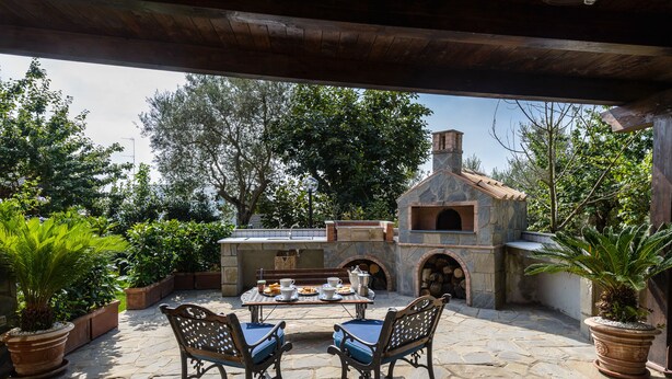 An outdoor patio with stone flooring, two wooden chairs, a table set with a teapot, cups, and a plate of food, and a stone oven in the background.