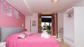 Een slaapkamer met een roze sprei, een witte nachtkastje, een houten stoel en uitzicht op een terras met een tafel en stoelen.