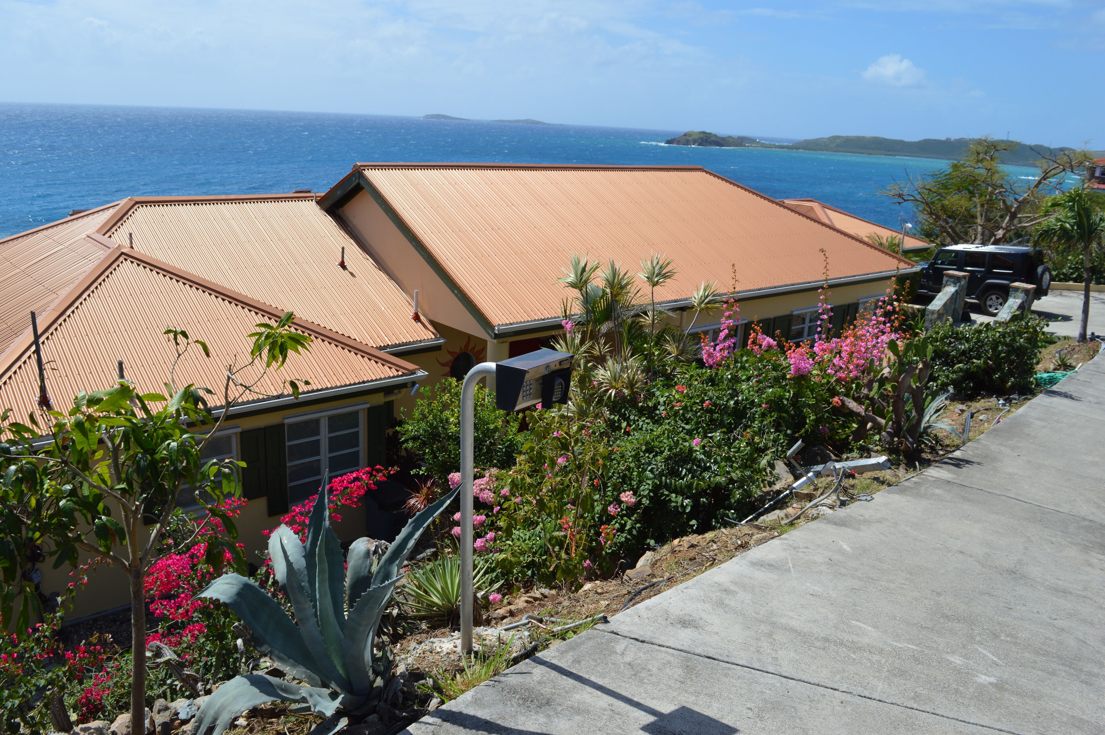 Une propriété située sur la côte, avec une grande maison dotée d’un toit en tuiles, d’une allée pavée et d’un aménagement paysager luxuriant comprenant des fleurs roses et des plantes tropicales.