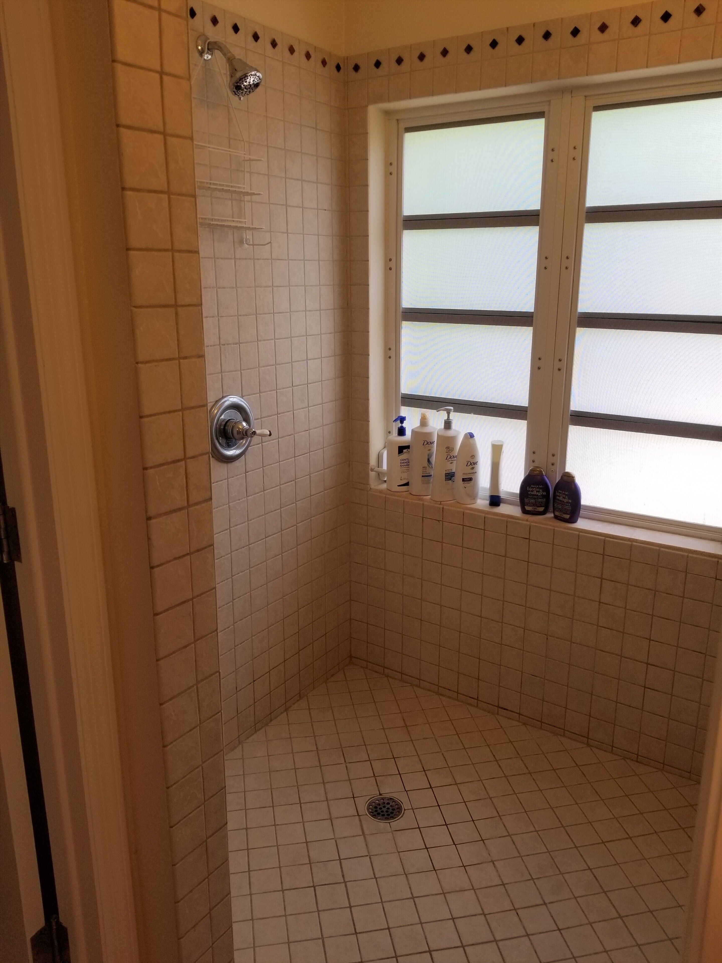 Une petite salle de douche carrelée, avec une fenêtre et des articles de toilette posés sur le rebord.