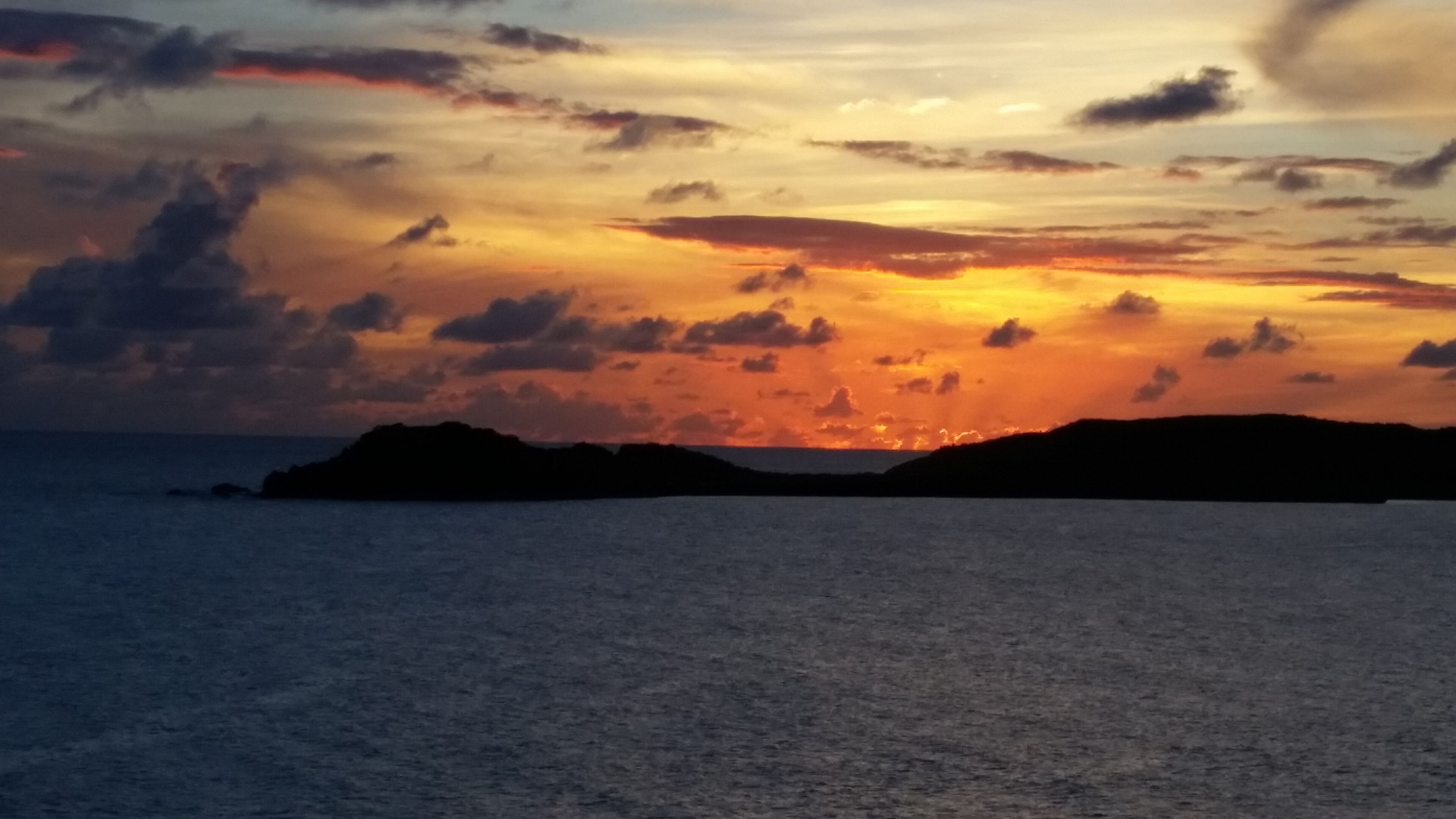 Solnedgang ved kysten med silhuetter af land og et roligt hav.