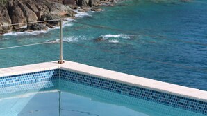 En swimmingpool med kristallklart blått vatten, omgiven av en klippig kust och frodig grönska.